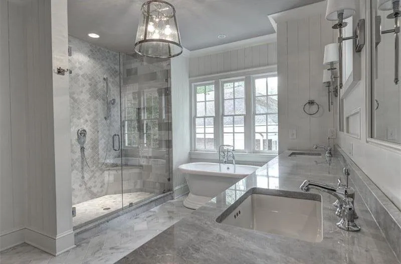 A luxurious bathroom featuring marble countertops, white walls, a freestanding tub, glass shower enclosure, and a large window.